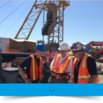 three male workers at a construction site with a crane in the background all three are wearing hard hats and orange safety vests